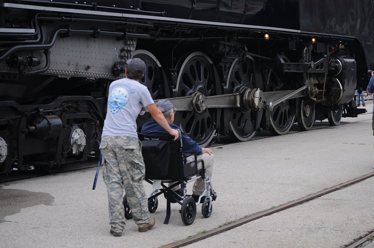 grandson pushing grandfather in wheelchair next to steam locomotive